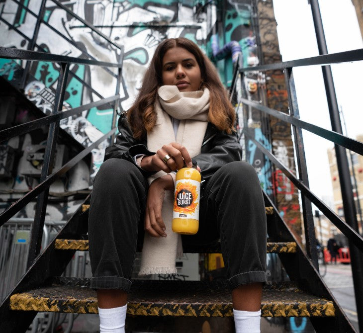A girl is sitting on some metal steps holding a bottle of JUICEBURST Orange Juice, behind her there is a wall of graffiti